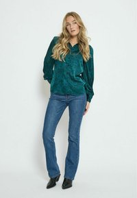 Grün gemusterte Bluse mit Knopfverschluss, Puffärmel, kombiniert mit enganliegenden blauen Jeans und schwarzen Ankle Boots. Glatte Stofftextur.