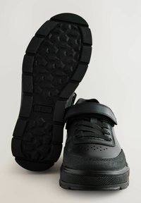 Sorte sportssko med en glat læderoverdel, mesh-detaljer, snørelukning og en velcro-rem. Gummisålen har et hexagonalt dækmønster.