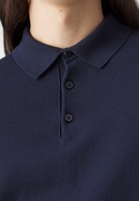 Gros plan d'une personne portant un polo bleu marine à boutons, avec trois boutons fermés au col.