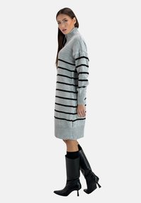 Robe en tricot grise avec des rayures horizontales noires, col haut et poignets côtelés. Portée avec des bottes noires à talons aiguilles hautes. Silhouette mince et élancée.