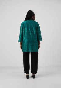 Teal kanten tuniek met bladmotief, driekwart mouwen en gegolfde zoom, gecombineerd met zwarte loszittende broeken en chunky haksandalen.