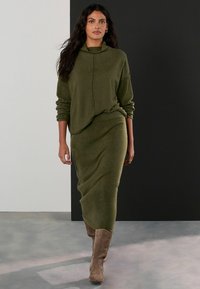 Olivew Greenski dwuczęściowy strój z luźną bluzą zapinaną na zamek i dopasowaną spódnicą do kostek, wykonany z teksturowanej dzianiny. Botki są jasnobrązowe.