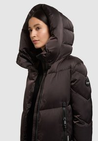 Dunkelbraune Steppjacke mit großer Kapuze, Reißverschluss und Druckknopfdetails. Verfügt über ein Logo-Patch auf der Schulter und eine gesteppten Textur.