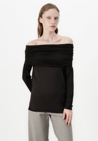 Black off-the-shoulder top with gathered fabric detail, long sleeves, and a smooth texture, paired with light gray trousers.