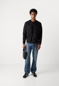 Jachetă bomber neagră cu două buzunare frontale, tricou negru, blugi bleumarin, teniși negri cu dungi albe, purtând o geantă neagră.