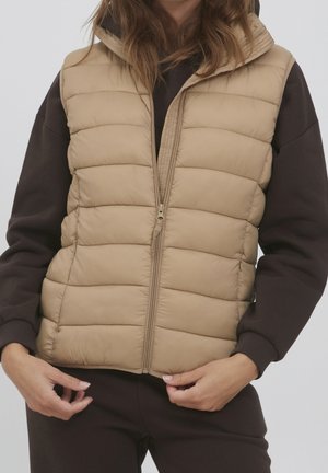 Person trägt eine beigen gesteppte ärmellose Weste über einem dunklen Langarmshirt und hält die unteren Ränder der Weste in der Nähe des Reißverschlusses fest.