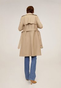 Osoba w beżowym płaszczu, niebieskich dżinsach i beżowych szpilkach, stojąca plecami do obiektywu na jednolitym tle.