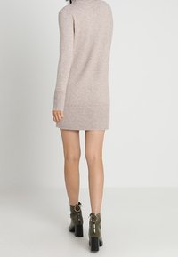 Femme portant une robe en maille beige clair à manches longues et des bottines à talons vert olive, debout devant un fond blanc uni.