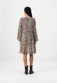 Woman with long braided hair wearing a knee-length leopard print dress and black ankle boots, standing against a plain white background.