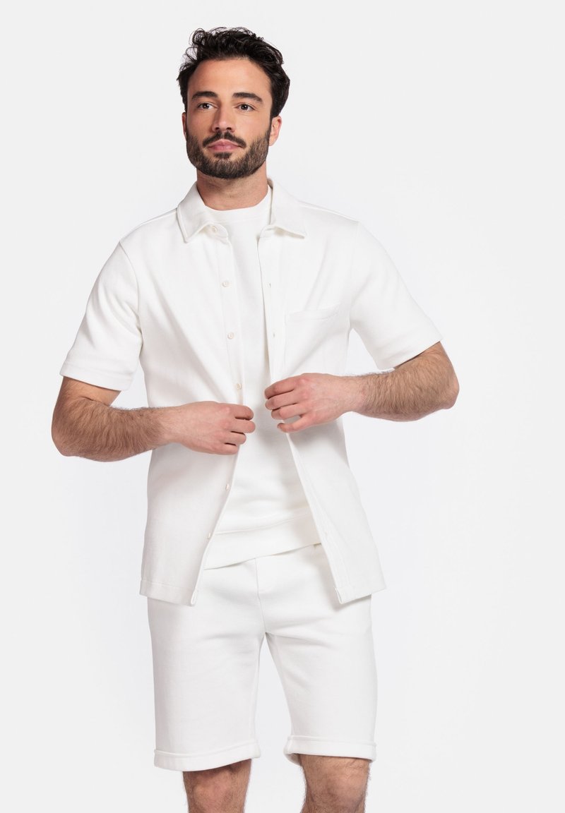 Witte short-sleeve shirt met knoopsluiting aan de voorkant, gelaagd over een bijpassend wit T-shirt met ronde hals en shorts, wat een strakke, monochrome uitstraling toont.