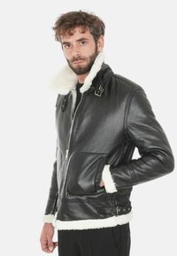 Giubbotto di pelle nera con colletto e polsini in pelliccia bianca, chiusura con zip sul davanti, tasche laterali e dettagli con fibbia in vita. Texture liscia.