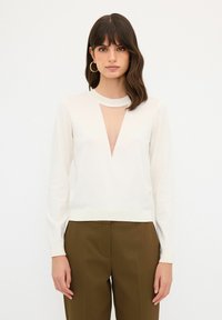 Weißer Pullover mit tiefem V-Ausschnitt am Hals und langen Ärmeln. Glatte Strickstruktur, kombiniert mit hochtaillierten braunen Hosen.