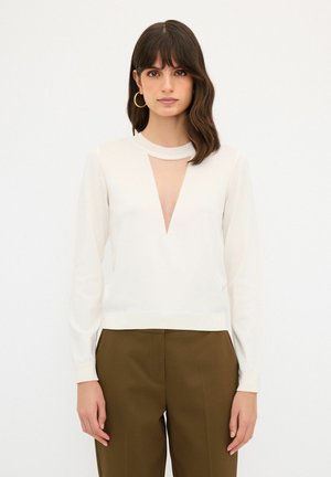 Weißer Pullover mit tiefem V-Ausschnitt am Hals und langen Ärmeln. Glatte Strickstruktur, kombiniert mit hochtaillierten braunen Hosen.