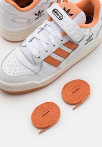 White leather sneakers with orange stripes and accents, textured details, and a Velcro strap; includes two orange shoelaces.