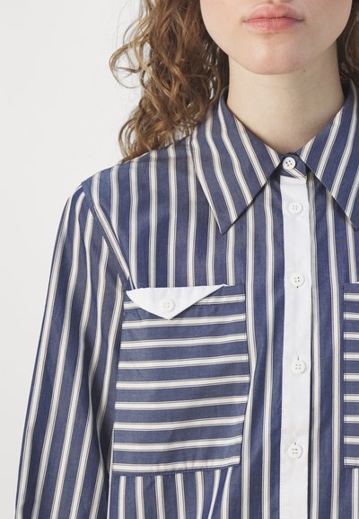 Camicia con bottoni, dotata di un ampio colletto, caratterizzata da una fantasia a righe verticali blu navy e bianche, con una tasca a toppa e una texture del tessuto liscia.