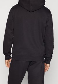 Sweat à capuche noir avec une texture lisse, des poignets et un ourlet côtelés, et une coupe décontractée. Présenté de dos, montrant des détails de couture minimaux.