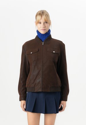 Kvinne med brun zip-up jakke over en blå polo genser og en marineblå plissé skjørt foran en plain hvit bakgrunn.