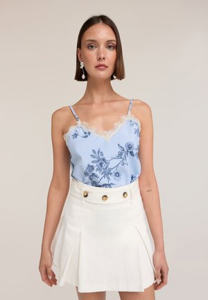 Femme portant un caraco bleu à fleurs orné de dentelle et une jupe plissée blanche taille haute avec des boutons dorés.