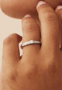 Bague en argent avec une pierre centrale transparente, présentant un anneau lisse et de petites pierres incrustées le long de l'anneau, sur un fond de teint.