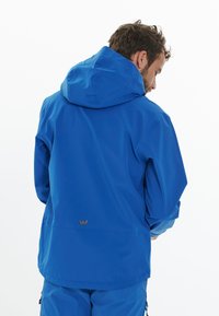 Sininen vedenpitävä takki hupulla, jossa on sileä pinta ja säädettävä helma. Takana on merkittävä ruskea brändilogo.