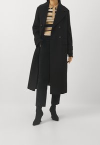 Manteau noir à double boutonnage avec grands revers, poches latérales et poignets retroussés, porté sur un haut rayé et un pantalon noir, associé à des bottes pointues.