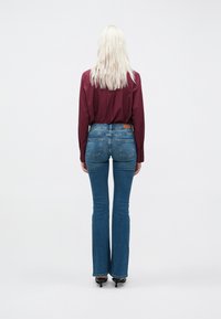 Jeans in denim blu scuro slim-fit con una leggera svasatura in fondo, accentuati da una toppa in pelle marrone sul retro, indossati con una camicia bordeaux.
