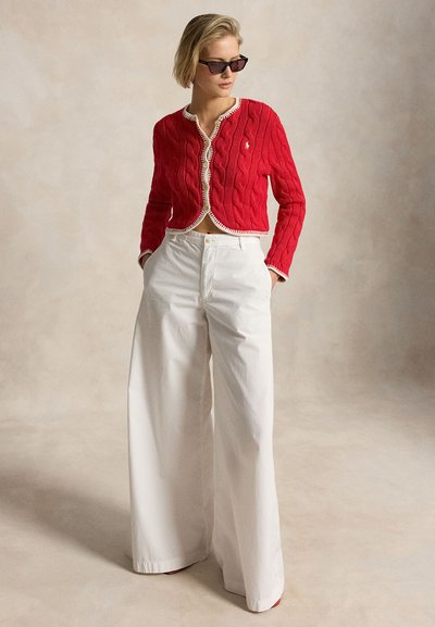Kobieta w czarnych okularach przeciwsłonecznych, czerwonym, krótkim swetrze na guziki z kremowym wykończeniem oraz białych, szerokich spodniach z wysokim stanem, stoi z rękami w kieszeniach.