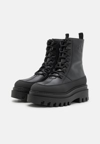 Černé platformové bojové boty s gumovou podrážkou, šněrováním v přední části, texturedními panely a vyraženým logem na boku.