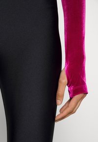 Mustat legginsit, joissa on sileä pinta, yhdistettynä pitkähiharaiseen, kirkkaan vaaleanpunaisesta sametista valmistettuun puseroon, jossa on tiukka istuvuus ja peukalonaukko.