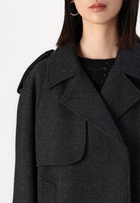 Manteau en mélange de laine gris foncé avec un col cranté, des revers oversize et des poches latérales. Le tissu présente une texture subtile avec une coupe ajustée.