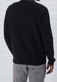 Felpa nera con maniche raglan, scollatura e orlo a coste, realizzata in tessuto morbido. Indossata con jeans grigi chiari, mostrando la vista posteriore.