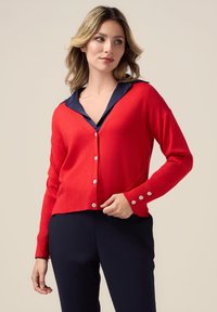 Cardigan rouge avec un col en V, des manches longues et des accents de boutons argentés. Présentant un col superposé marine et un design court. Porté sur un pantalon marine.