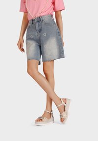 T-shirt rosa chiaro con perlini, shorts di jeans a vita alta con decorazioni a forma di cuore e sandali platform beige con cinturini intrecciati.