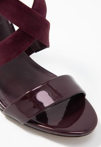 Bordeaux vysoké sandály na podpatku s lesklým lakovaným prstovým páskem a měkkým zamší křížovým páskem kolem kotníku. Hladká textura; otevřený design na špičce.