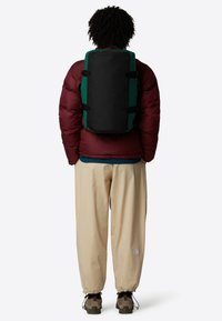 Black and green backpack with a rectangular shape, adjustable straps, and durable material. Worn over a maroon puffer jacket and beige pants.