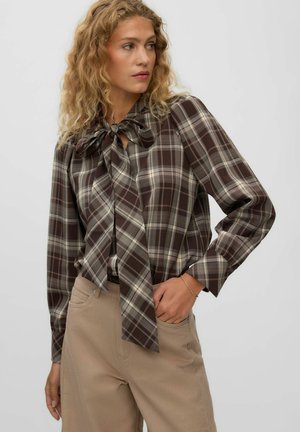 Femme aux cheveux blonds bouclés portant un chemisier à carreaux marron avec un grand nœud au col et un pantalon beige sur fond gris uni.