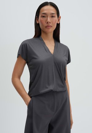 Frau mit langen, dunklen Haaren, die ein dunkelgraues, kurzärmeliges V-Ausschnitt-Oberteil und passende Hosen trägt, steht mit einer Hand in der Tasche.