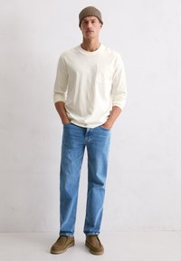 Mann mit beigem Beanie, weißem Langarmshirt mit Brusttasche, blauen Jeans und braunen Schuhen, der vor einer schlichten weißen Wand steht.