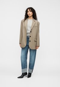 Μπεζ oversized σακάκι με τέσσερα κουμπιά, φορεμένο πάνω από γκρι πουλόβερ και μπλε τζιν με φθαρμένα τελειώματα, συνδυασμένο με μαύρα μυτερά μποτάκια αστραγάλου.