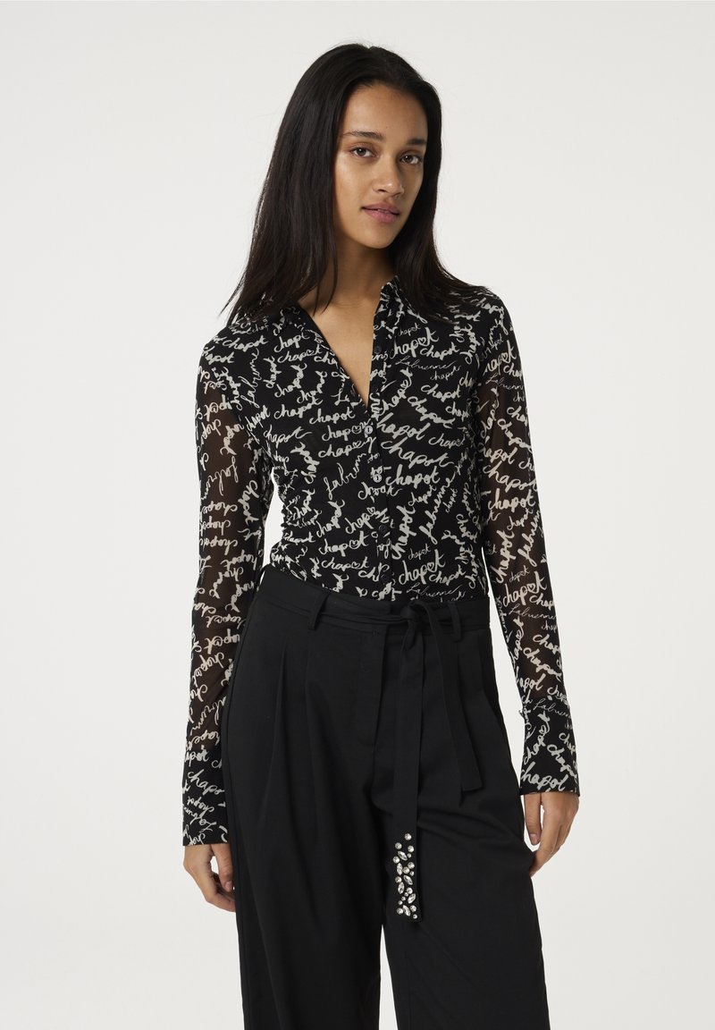 Zwarte blouse met lange mouwen van doorschijnende stof en een patroon van met de hand geschreven witte tekst. Heeft een kraag en een gebogen zoom.
