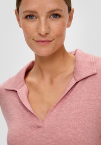 Jasnoróżowy sweter polo o miękkiej teksturze, z kołnierzykiem i dekoldem w kształcie litery V. Tkanina wydaje się lekka i casualowa.