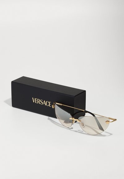 Aurinkolasit kultakehyksillä, kissamaisella muodolla, harmailla linsseillä ja mustilla yksityiskohdilla, esillä mustan Versace-kotelon vieressä.
