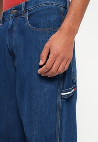 Σκούρο μπλε τζιν παντελόνι με κλασική γραμμή, που διαθέτει αντίθετο ράψιμο, μπροστινή τσέπη και πλάγια τσέπη με κόκκινη, λευκή και μπλε ρίγα.