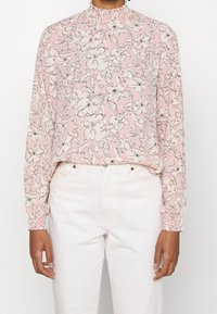 Ljusrosa blus med långärmar, vit blommönster, volangkrage och elastiska manschetter, parat med vita byxor.