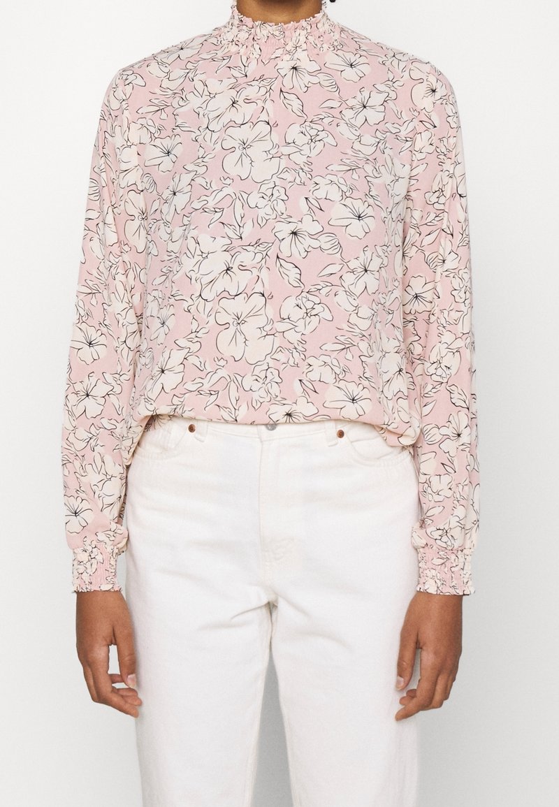 Ljusrosa blus med långärmar, vit blommönster, volangkrage och elastiska manschetter, parat med vita byxor.