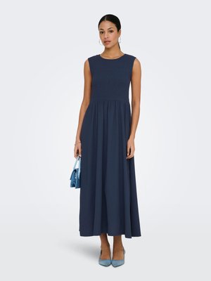 Donna che indossa un vestito midi blu navy senza maniche, tiene in mano una borsa azzurro chiaro e porta tacchi azzurro chiaro coordinati, in piedi davanti a uno sfondo uniforme.