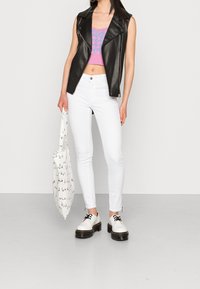 Mujer con chaleco de cuero negro, top corto morado, jeans ajustados blancos, zapatos de plataforma blancos, sosteniendo un bolso tote blanco con estampados negros.