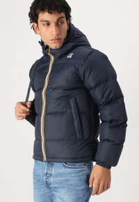 Giacca piumino blu navy con chiusura a zip e striscia gialla. Presenta un cappuccio, polsini elasticizzati e una tasca laterale. Materiale testurizzato.
