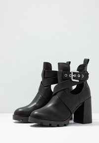 Botas de tobillo de cuero negro, de tacón alto y ancho, con diseño de correas en forma de cruz y detalle de hebilla plateada. Textura suave y puntera redonda.