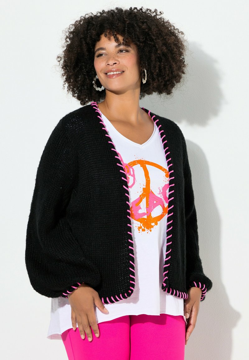 Schwarze Strickjacke mit pinkfarbenem Rand, lockere Passform, Ballonärmel. Getragen über einem weißen T-Shirt mit orange-pinkem Grafikdesign.